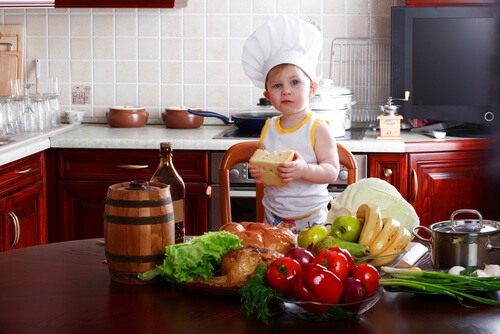 A toddler wearing a chef's hat, holding a block of cheese, and standing on a chair by a able full of vegetables.