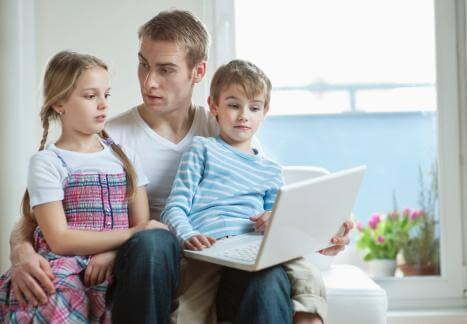 A father looking at a laptop computer with his 2 kids on his lap.