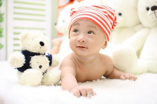 A baby wearing a striped cap lifting up his head while lying face down next to a teddy bear.