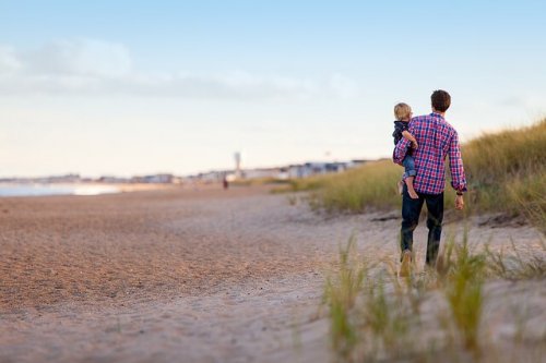 A father holding his toddler son as they walk along the beach.