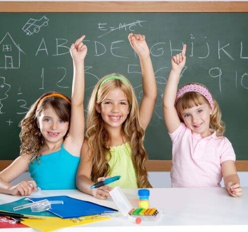 Three young girls in a classroom raising their hands.