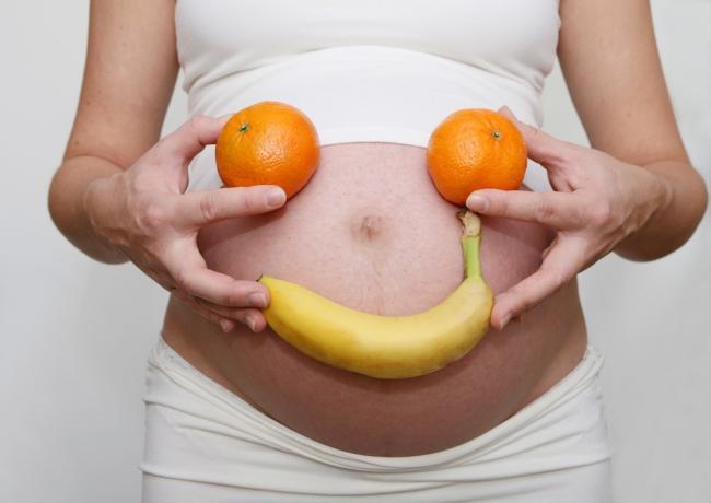 A woman holding a banana and two oranges in fronto of her belly, creating a smiley face along with her naval.