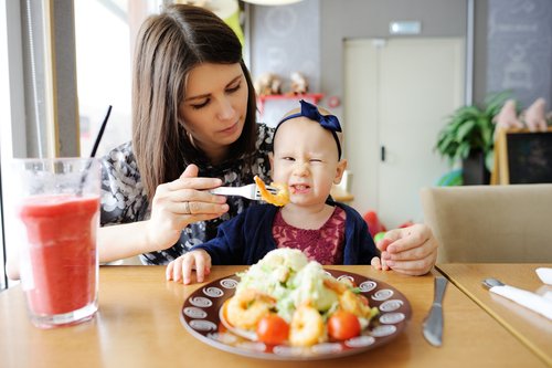 Mamá da de comer a su hija sentada sobre su regazo. A la niña no le gusta la comida que le dan.