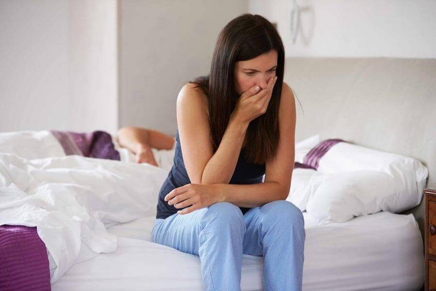 A woman sitting on the edge of her bed feeling nauseous.