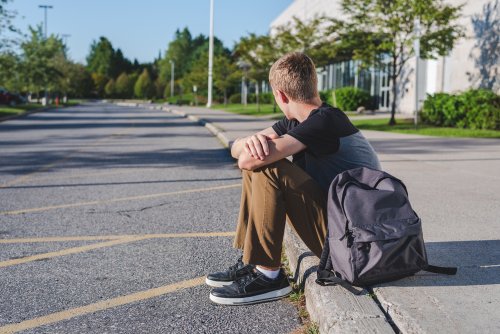 Chico adolescente sentado en el bordillo a la salida de la escuela porque no quiere estudiar.