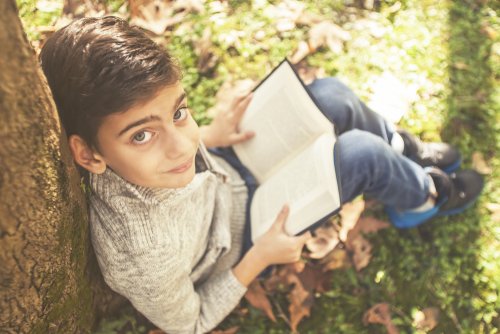 Niño leyendo sentado en un parque con hojas por el suelo.