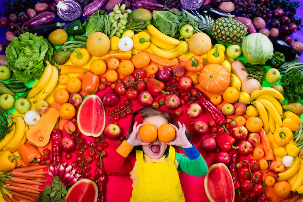Niña rodeada de un arcoíris de frutas y verduras.