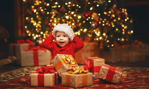 Bebé abriendo sus regalos de Navidad bajo el árbol decorado con luces.