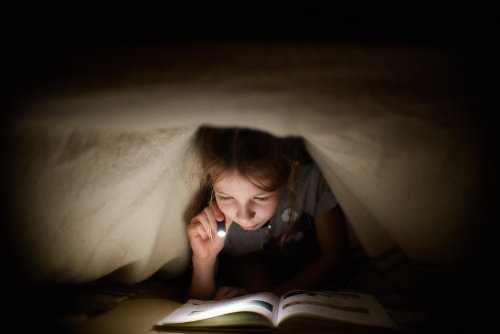 Niña leyendo uno de los cuentos de la colección ¡Mira debajo! bajo las sábanas de su cama con una linterna.
