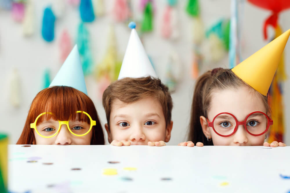 Children wearing birthday hats, peaking over a decorated table at a birthday party.