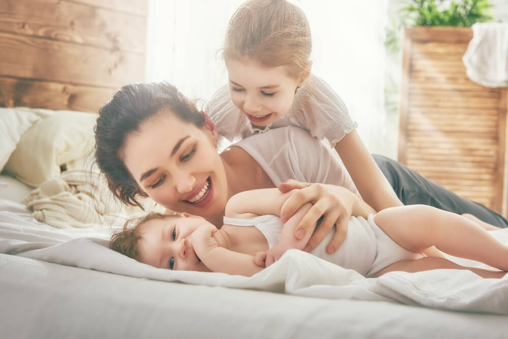 A mother playing in bed with her young daughter and her baby.