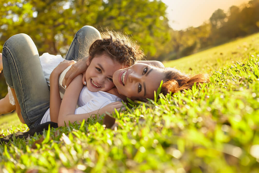 A mother lying on her back in the grass, hugging her little girl.