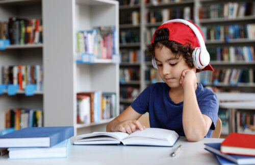 Niño leyendo un libro en la biblioteca con los auriculares puestos.