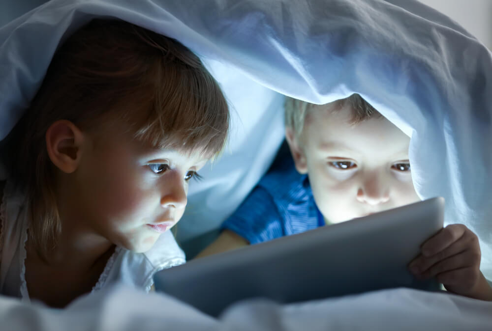 Des enfants regardent un film sous les couvertures sur une tablette.