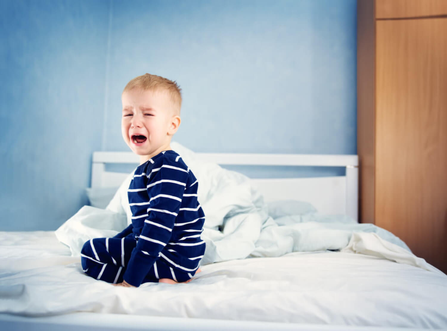 A toddler boy sitting on his bed crying.