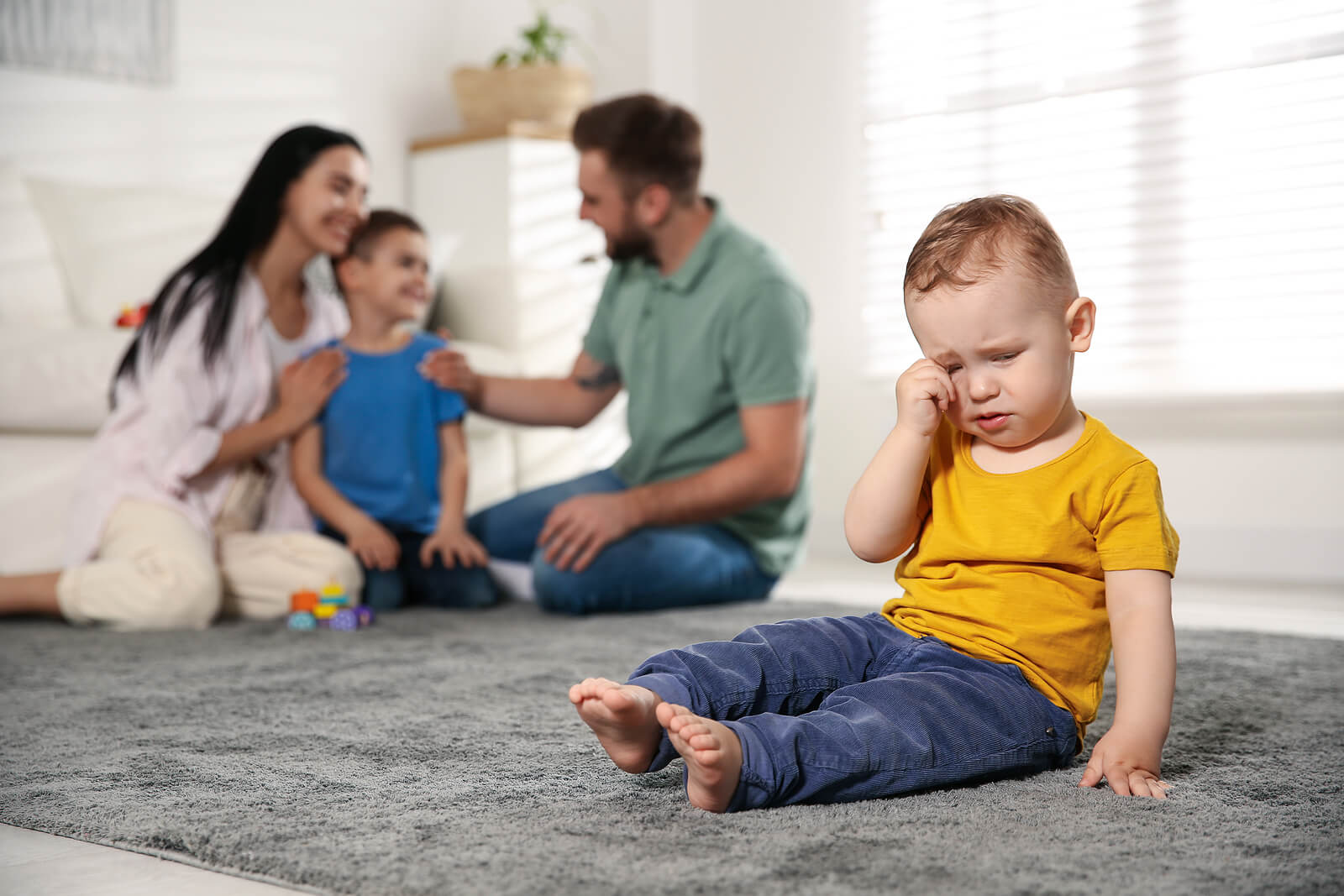 Hermano pequeño con celos llorando en el salón de casa mientras su padres adulan a su hermano mayor.