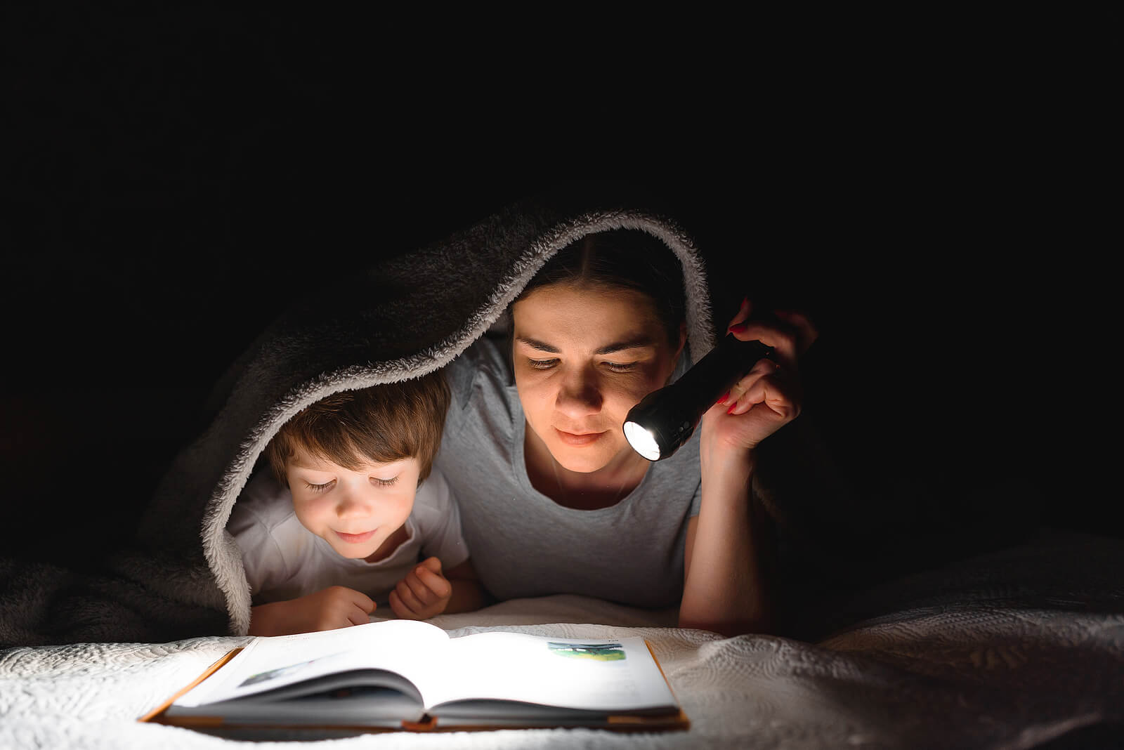 Madre con su hijo leyendo un cuento a la luz de la linterna mientras se tapan con las sábanas de la cama.