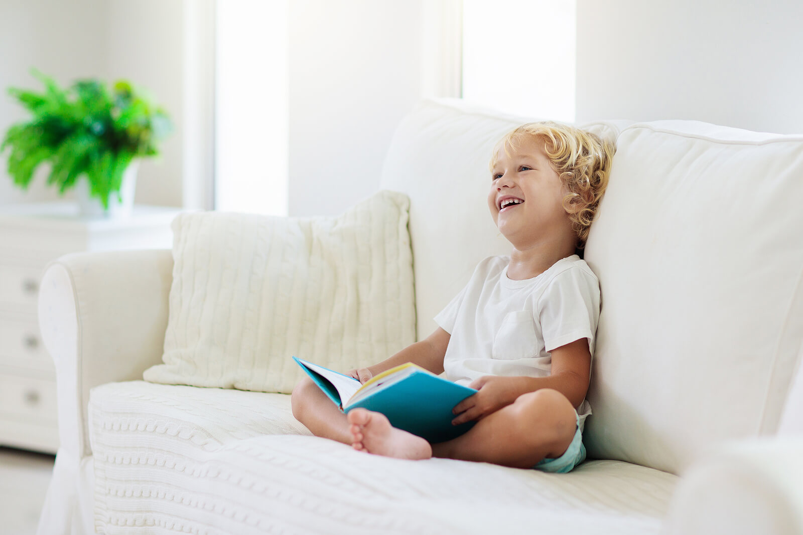 Un jeune garçon qui regarde un livre en souriant sur un canapé.