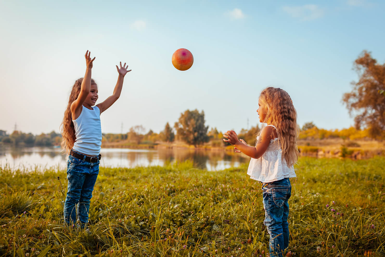 Two young girls playing catch in the grass near a lake.