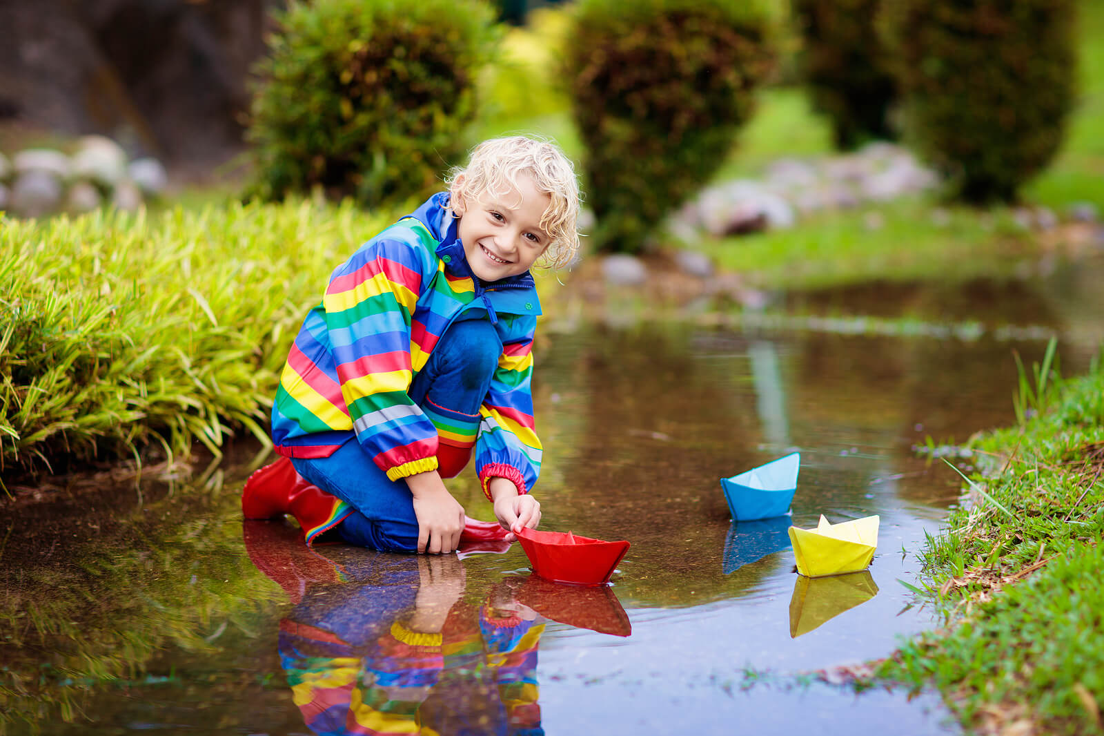 A child wearing a rain jacket while playing with paper boats in a stream.