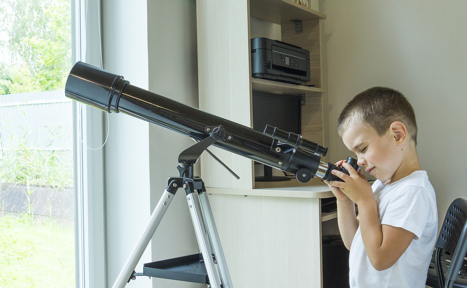En ung pojke tittar ut genom sitt fönster genom ett teleskop.