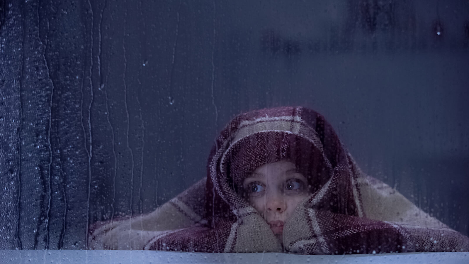 A child hiding under a blanket and looking out a window into the night rain.