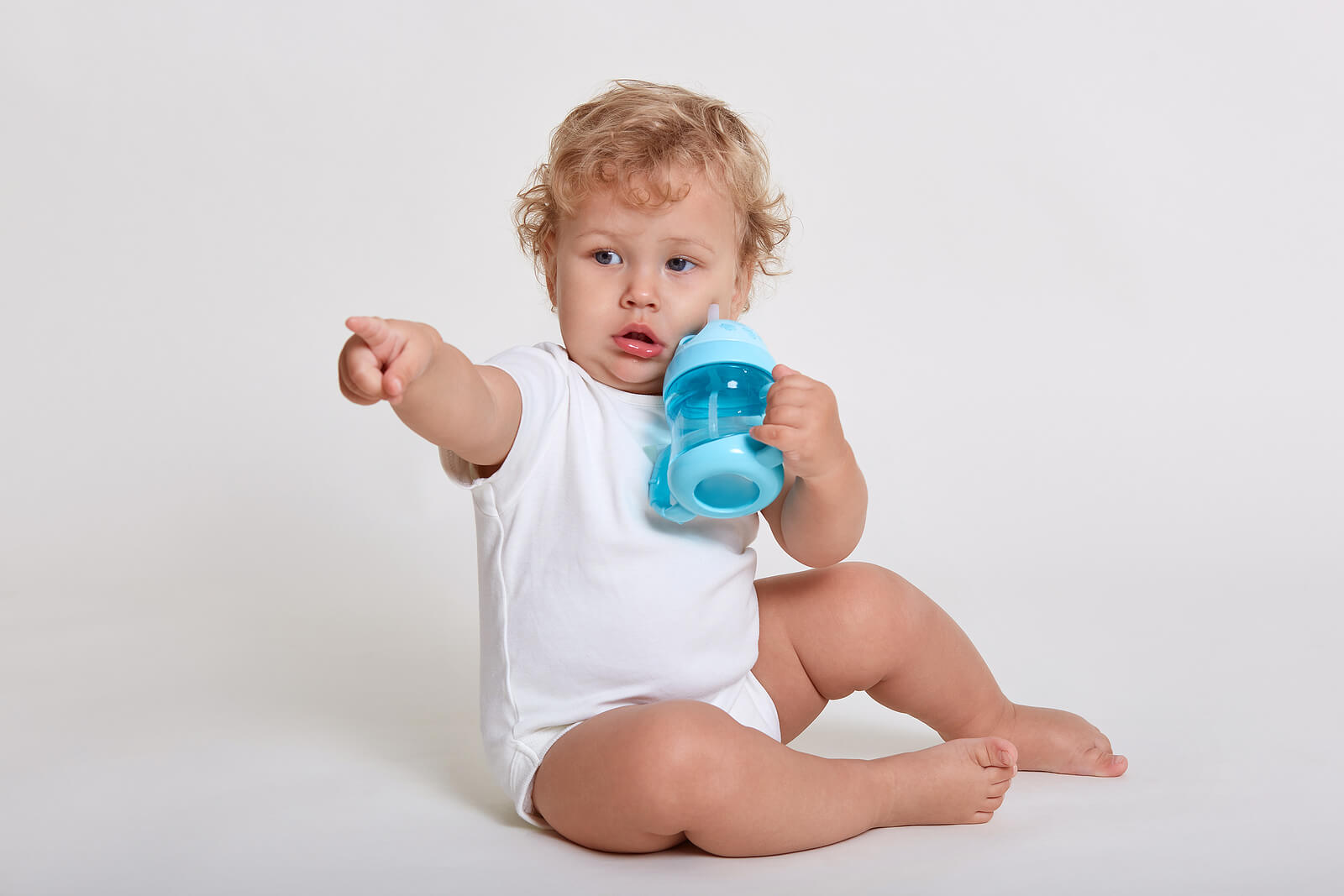 A baby holding a sippy cup and pointing.