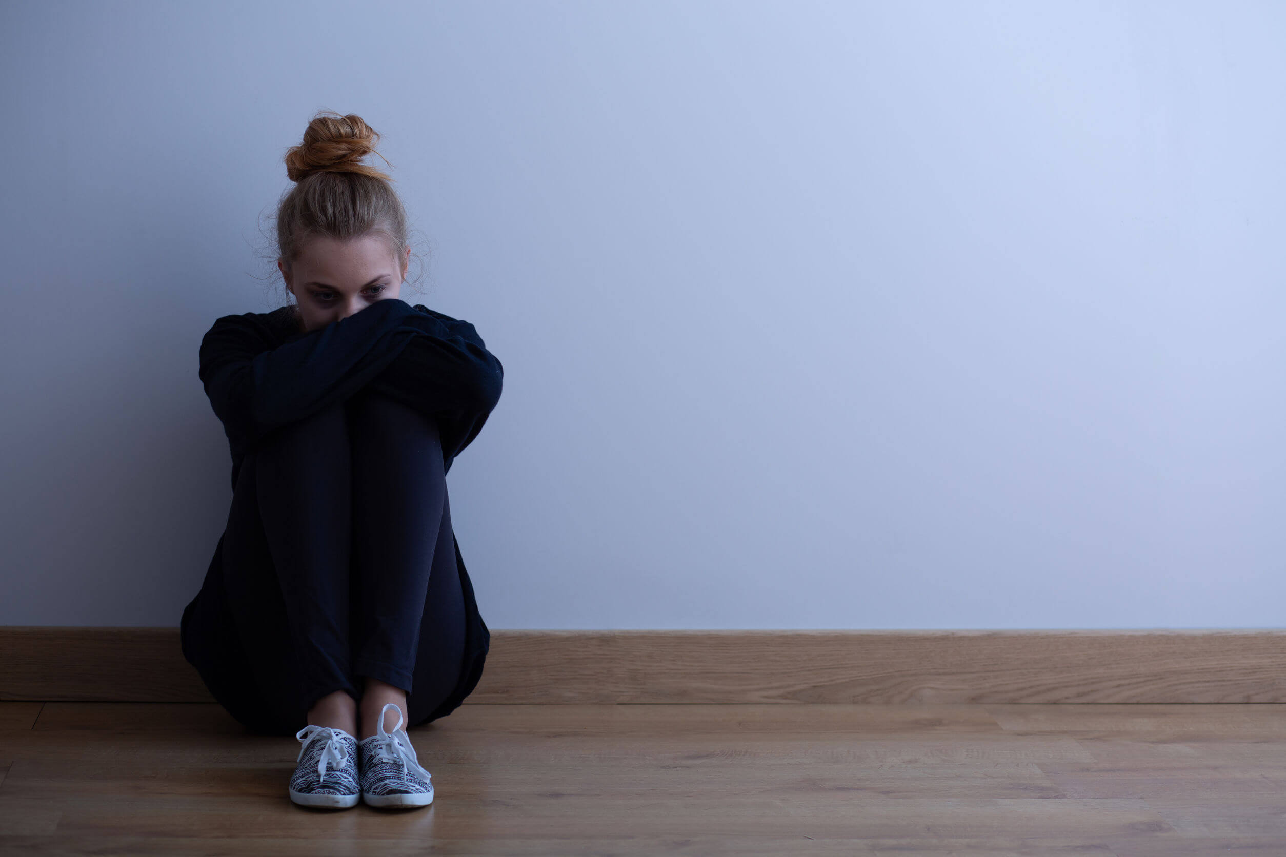 Mustaan pukeutunut teini-ikäinen tyttö istuu surullisena seinää vasten.