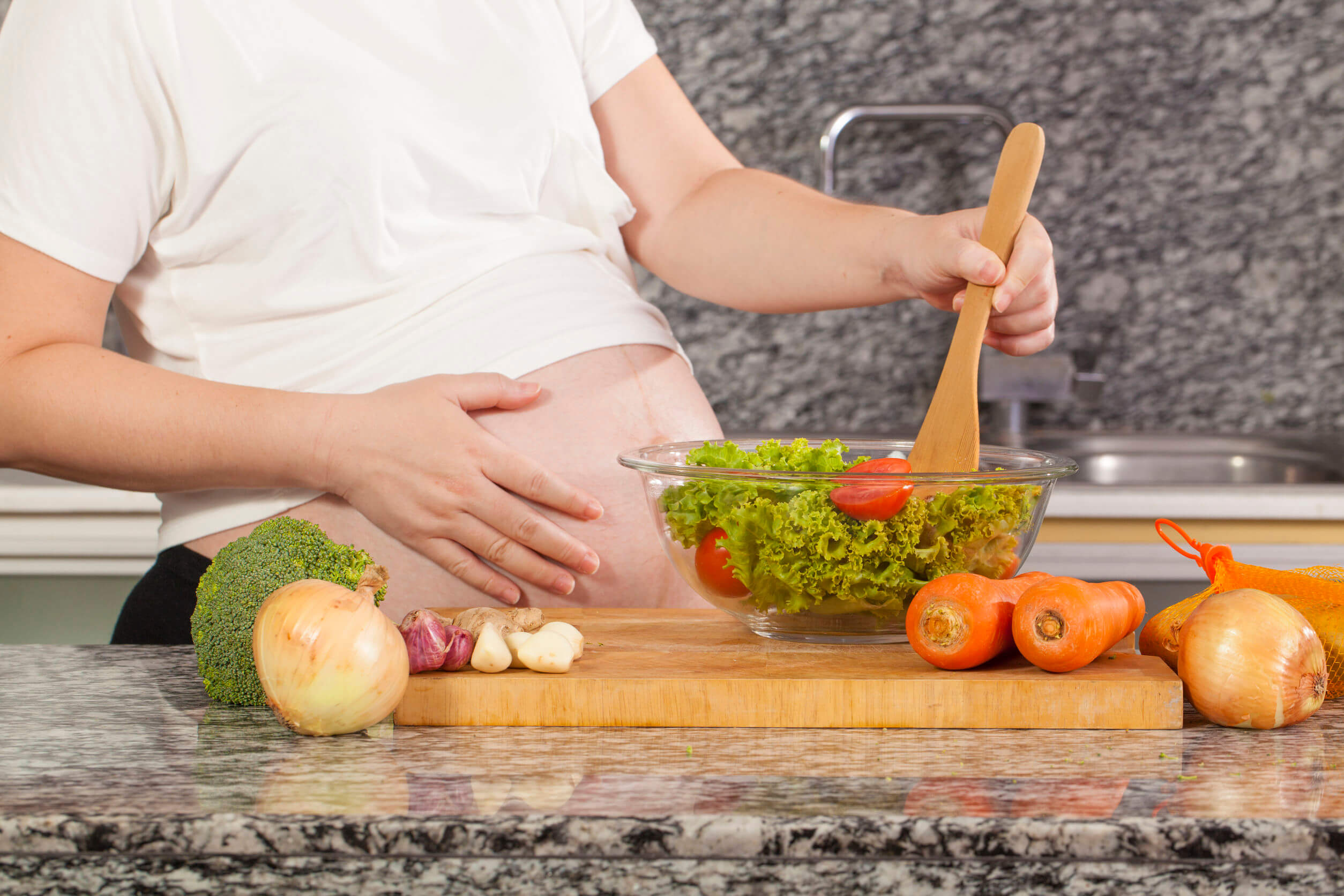 Mujer embarazada haciéndose una ensalada con verduras de hoja verde.