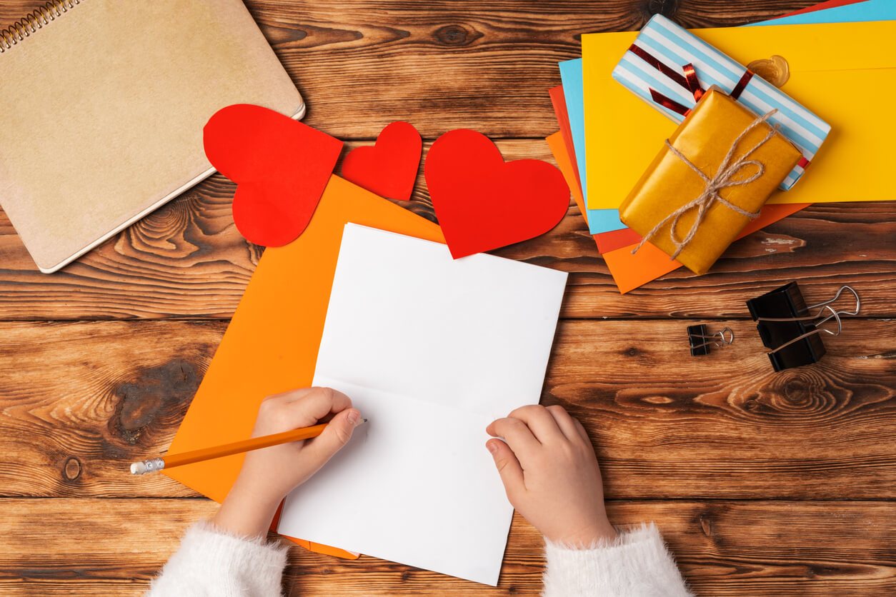 mano de niña escribiendo carta de amor en hoja de papel y corazones de colores sobre la mesa