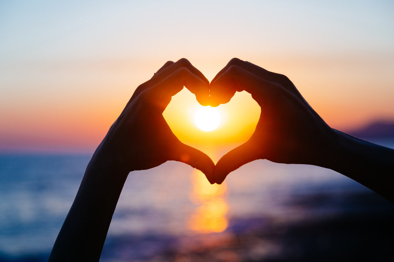 A woman making a heart with her hands with the sunset showing through.