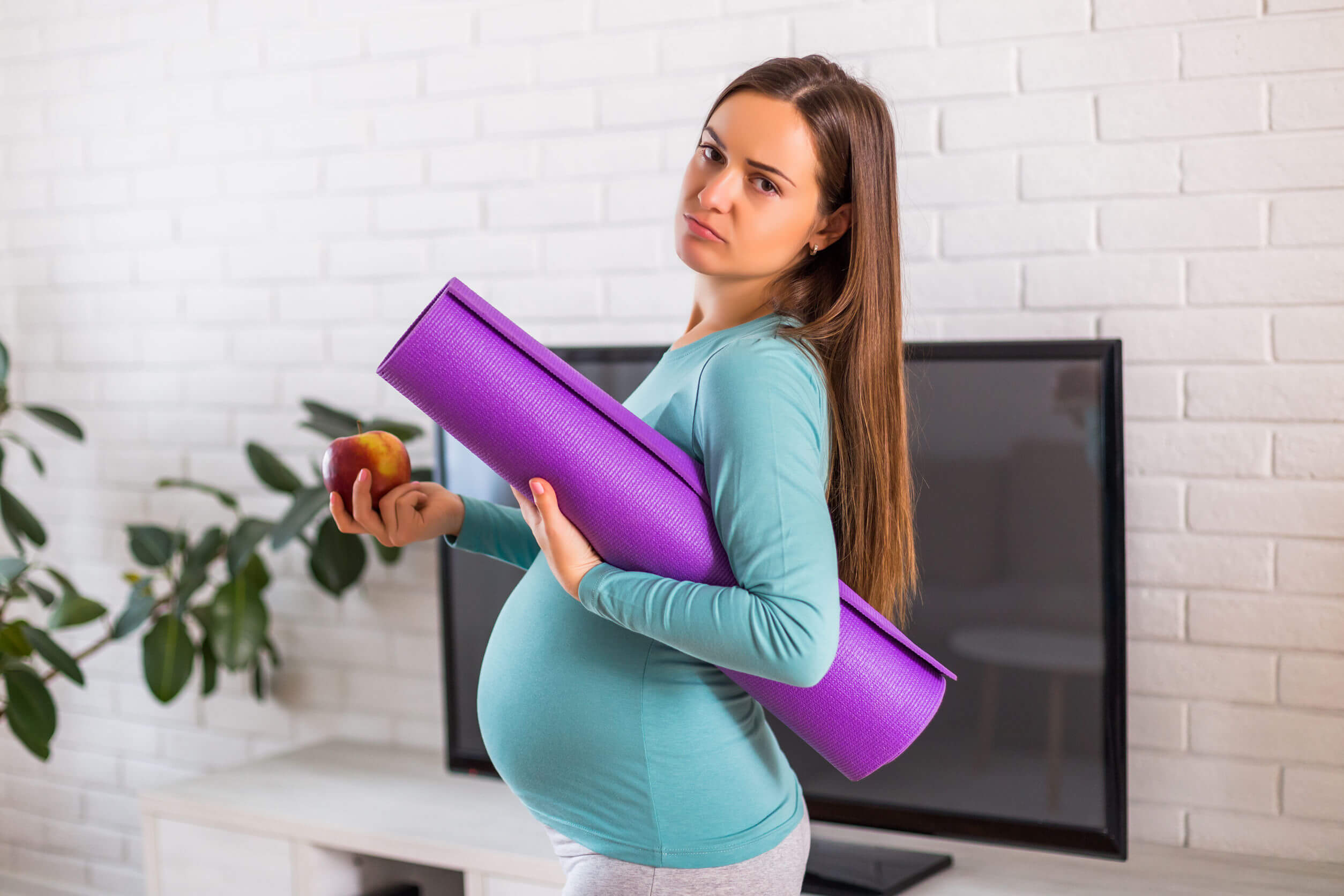 A pregnant woman holding an apple and carrying an exercise mat under her arm.