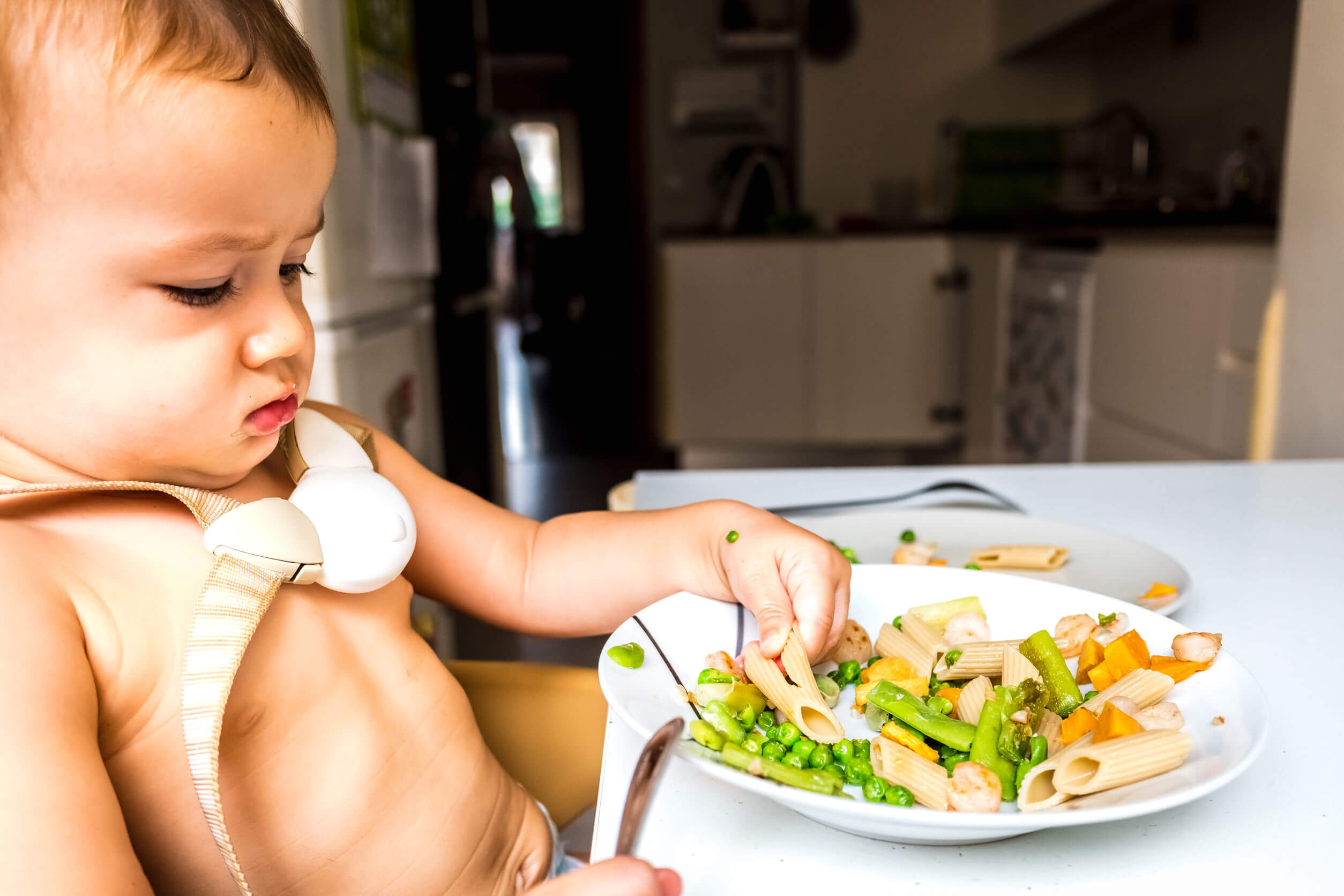 A baby sitting in a high chair looking at a plate of food skeptically.