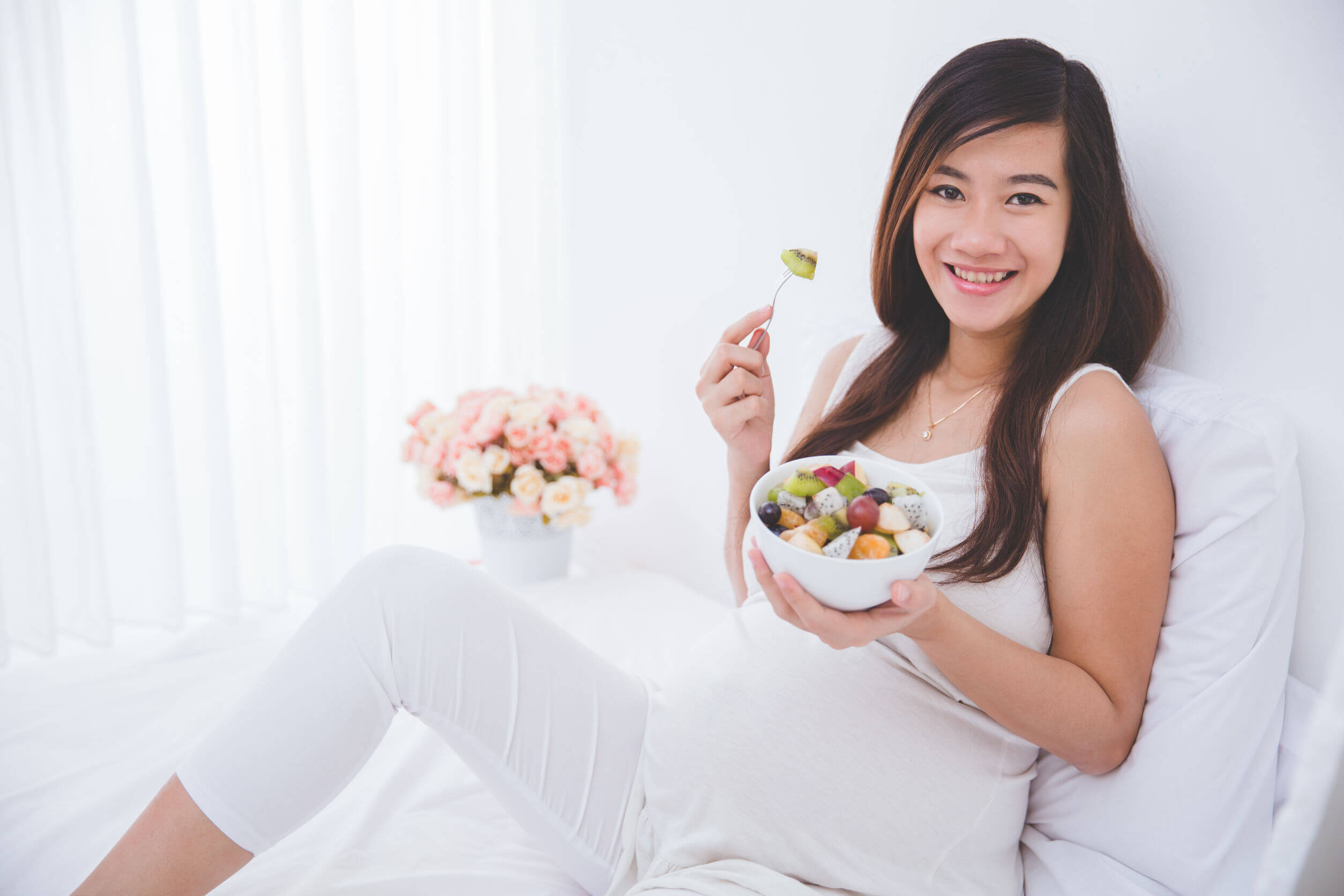 En gravid kvinna sitter i sängen och äter fruktsallad.