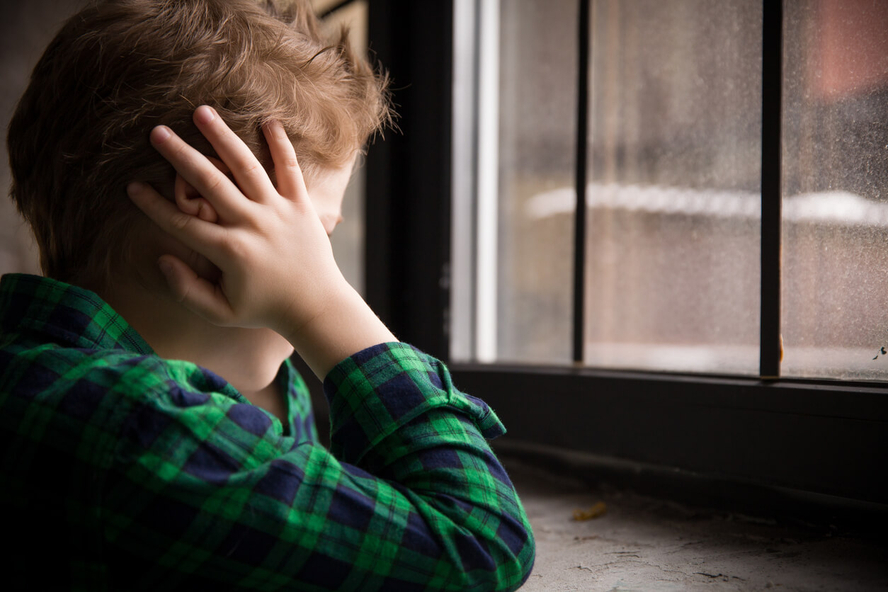 A child covering his ears and looking out a window.