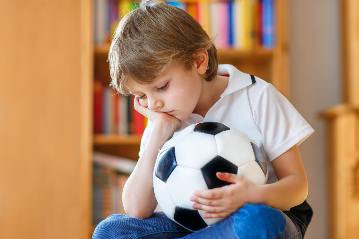 A sad child sitting indoors with a soccer ball in his lap.