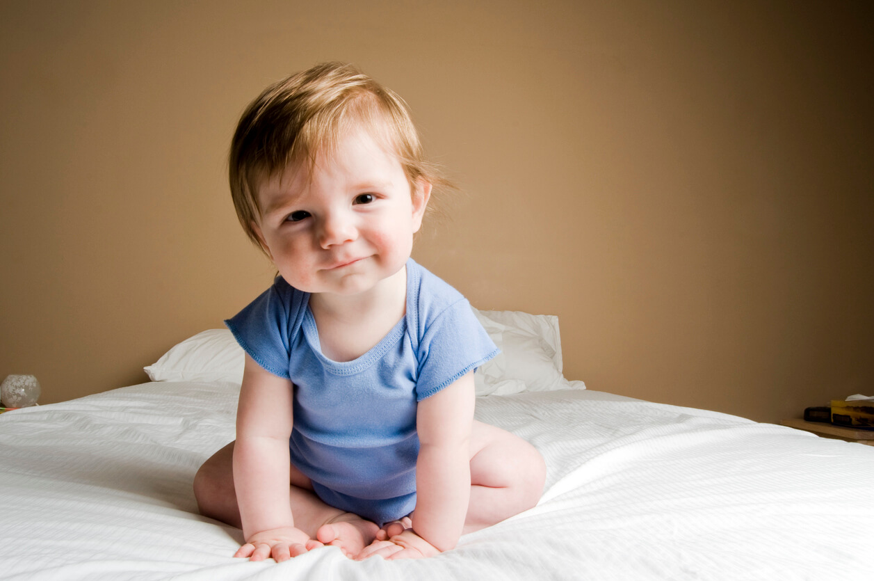 A baby boy sitting on a bed and leaning in to smile at the camera.