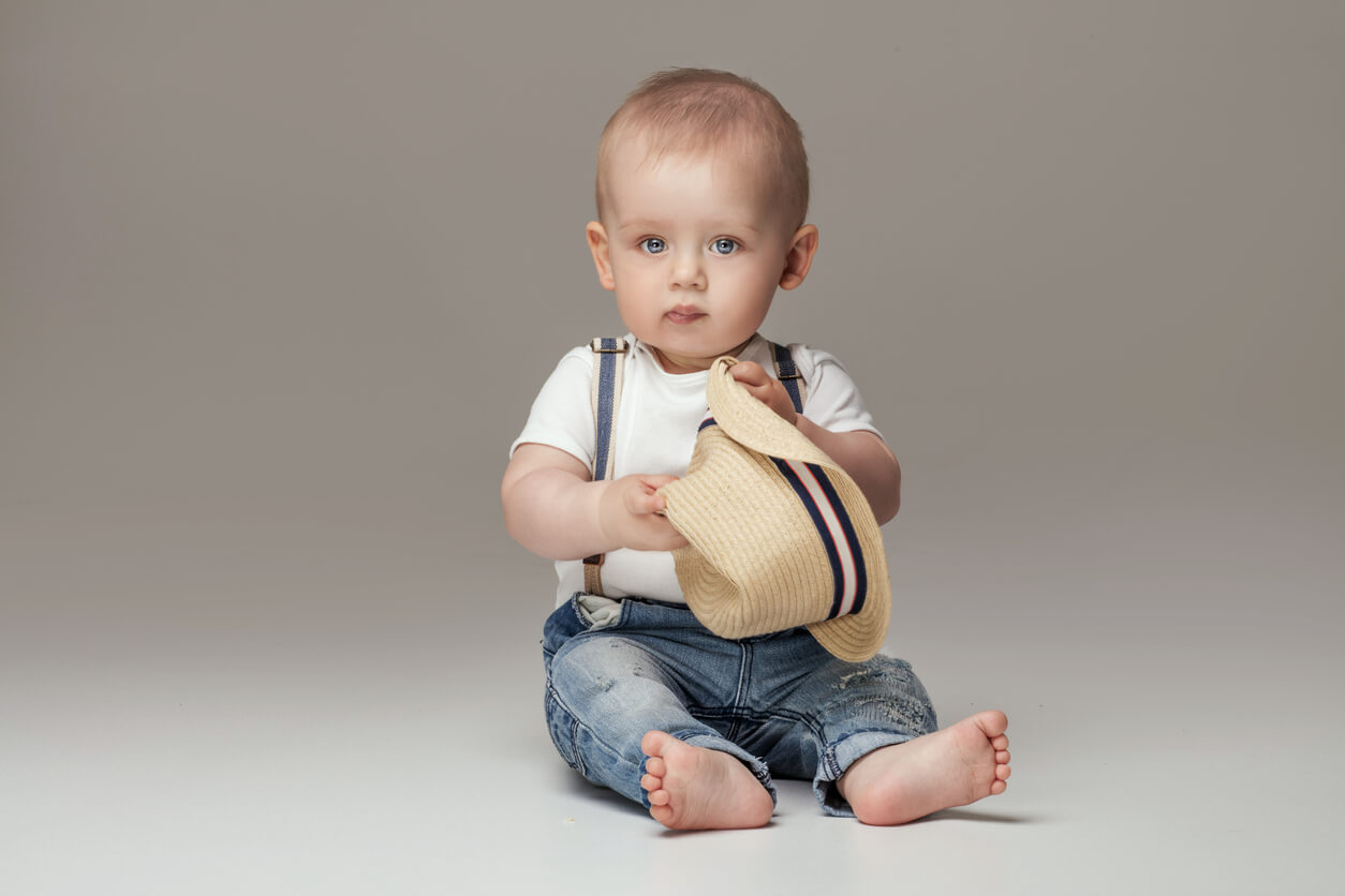 A baby boy wearing suspenders and holding a straw hat.