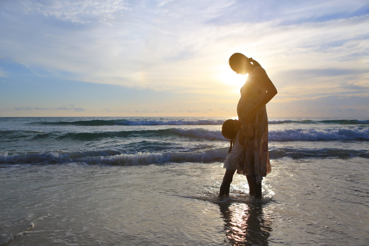 En liten jente som klemmer morens gravide mage mens de står ved kysten ved solnedgang.