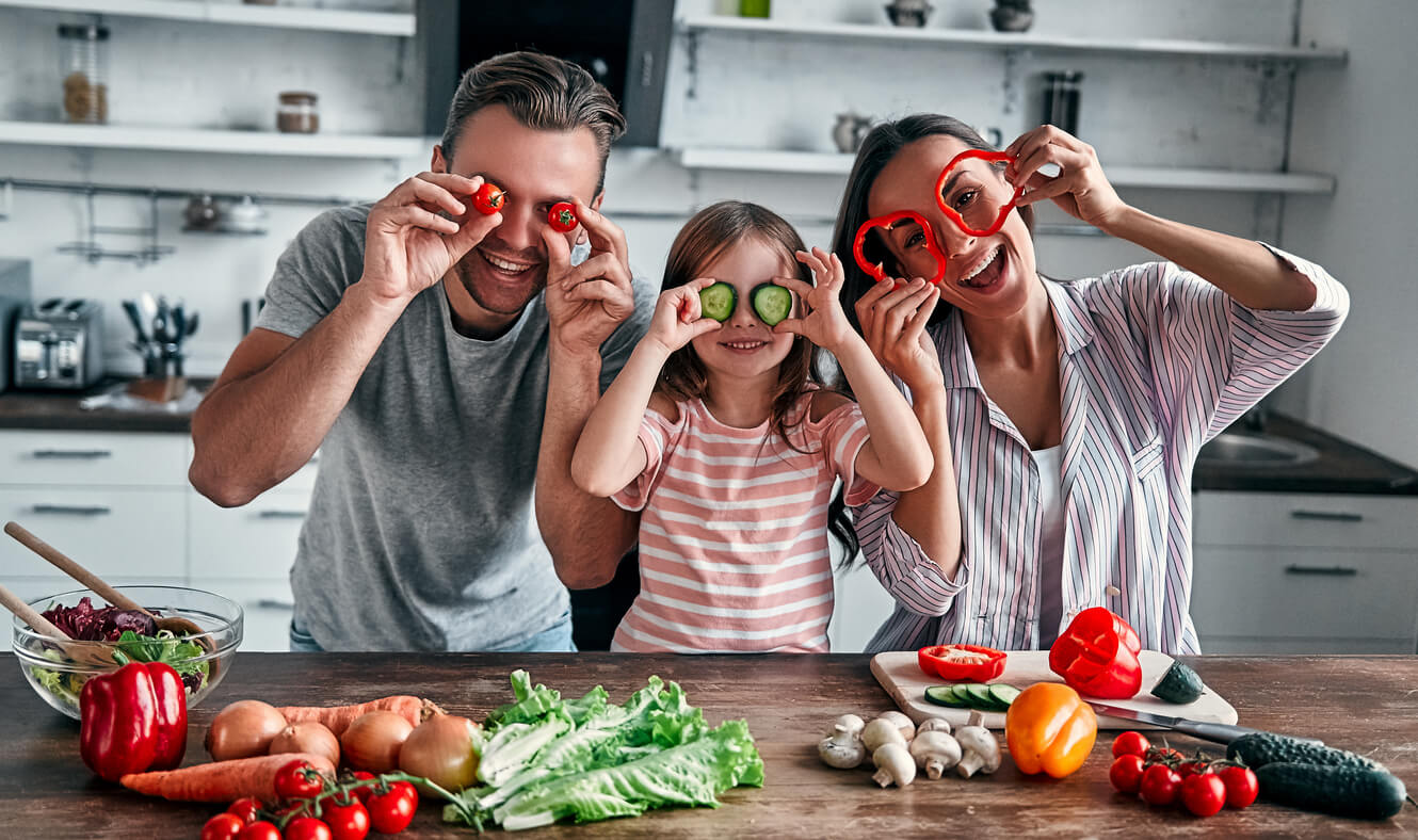 Padres con su hija en la cocina preparando frutas y verduras para proporcionar un buen aporte de vitaminas en la nutrición infantil.