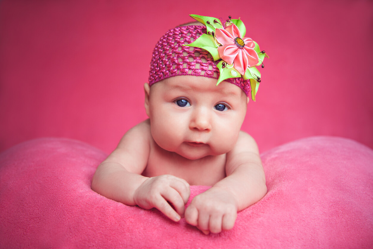 A baby wearing a pink knit cap leaning on a pink plush pillow.