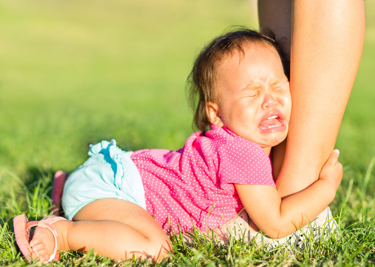 A baby crying on the ground and grabbing onto her mother's legs.