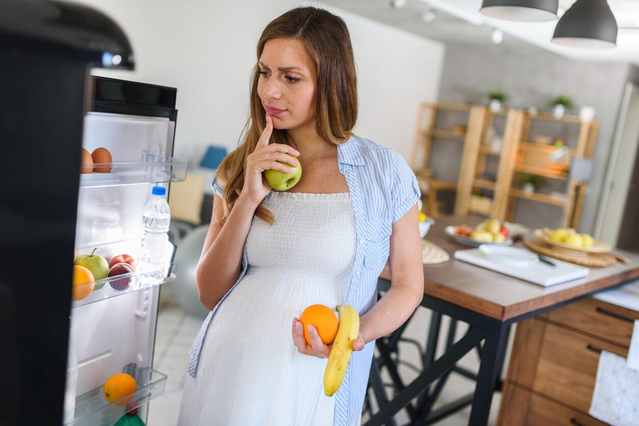 embarazada duda que come heladera puerta frutas verduras banana manzana naranja silla mesa