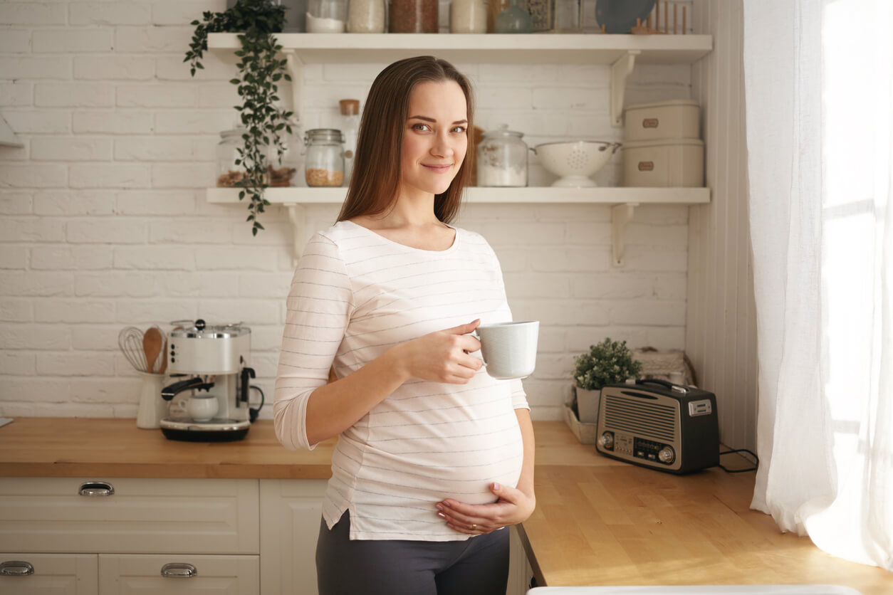 mujer embarazada sonrie dia mañana tarde toma cafe te taza cafetera cocina mesada panza feliz habito saludable