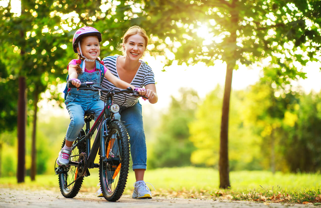 madre ensena nina nena montar pedalear bicicleta parque bosque feliz incentivo casco apoyo enseñanza aprendizaje crianza