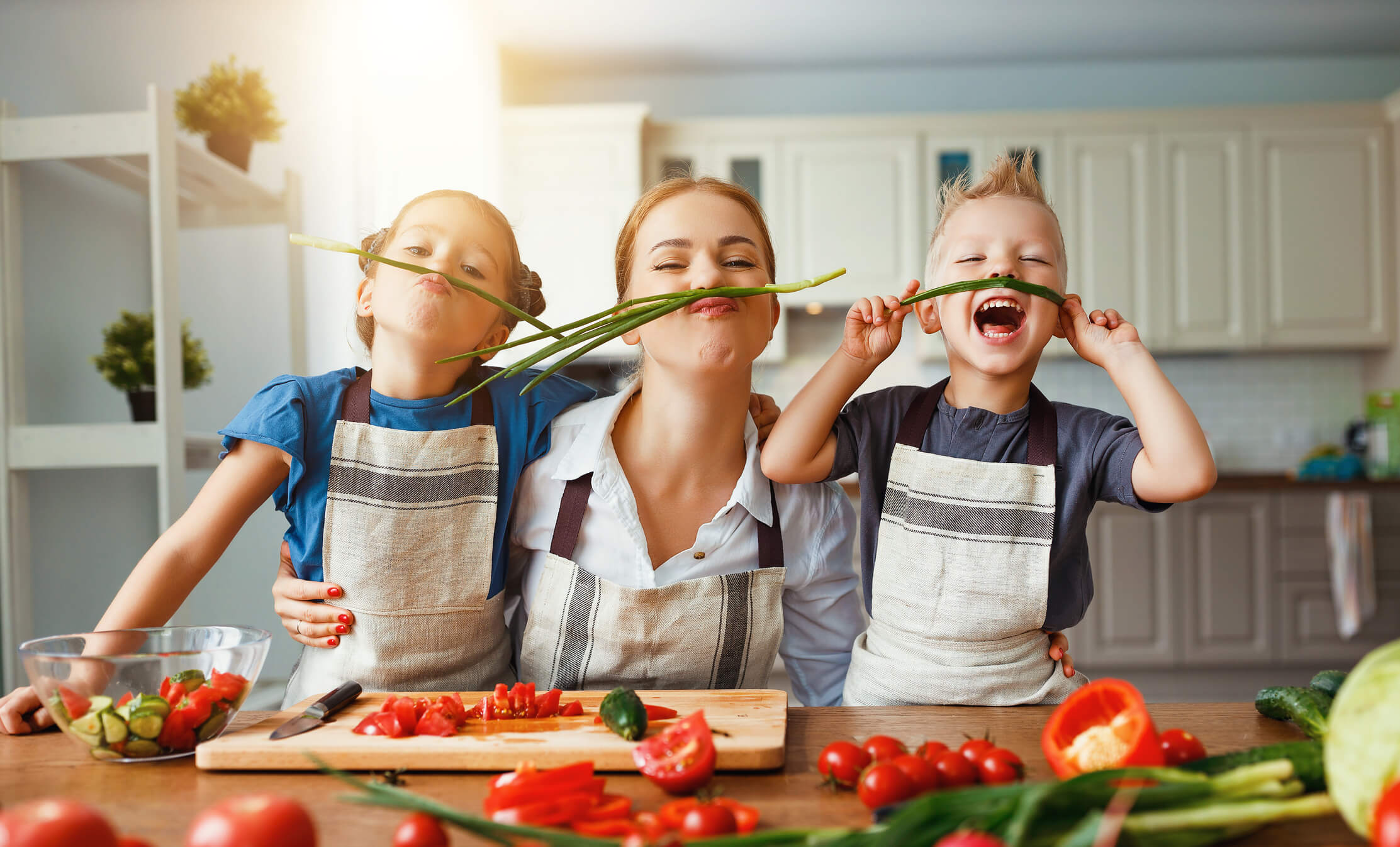 A mother and her 2 children wearing spring onions as mustaches while they prepare gazpacho recipes.