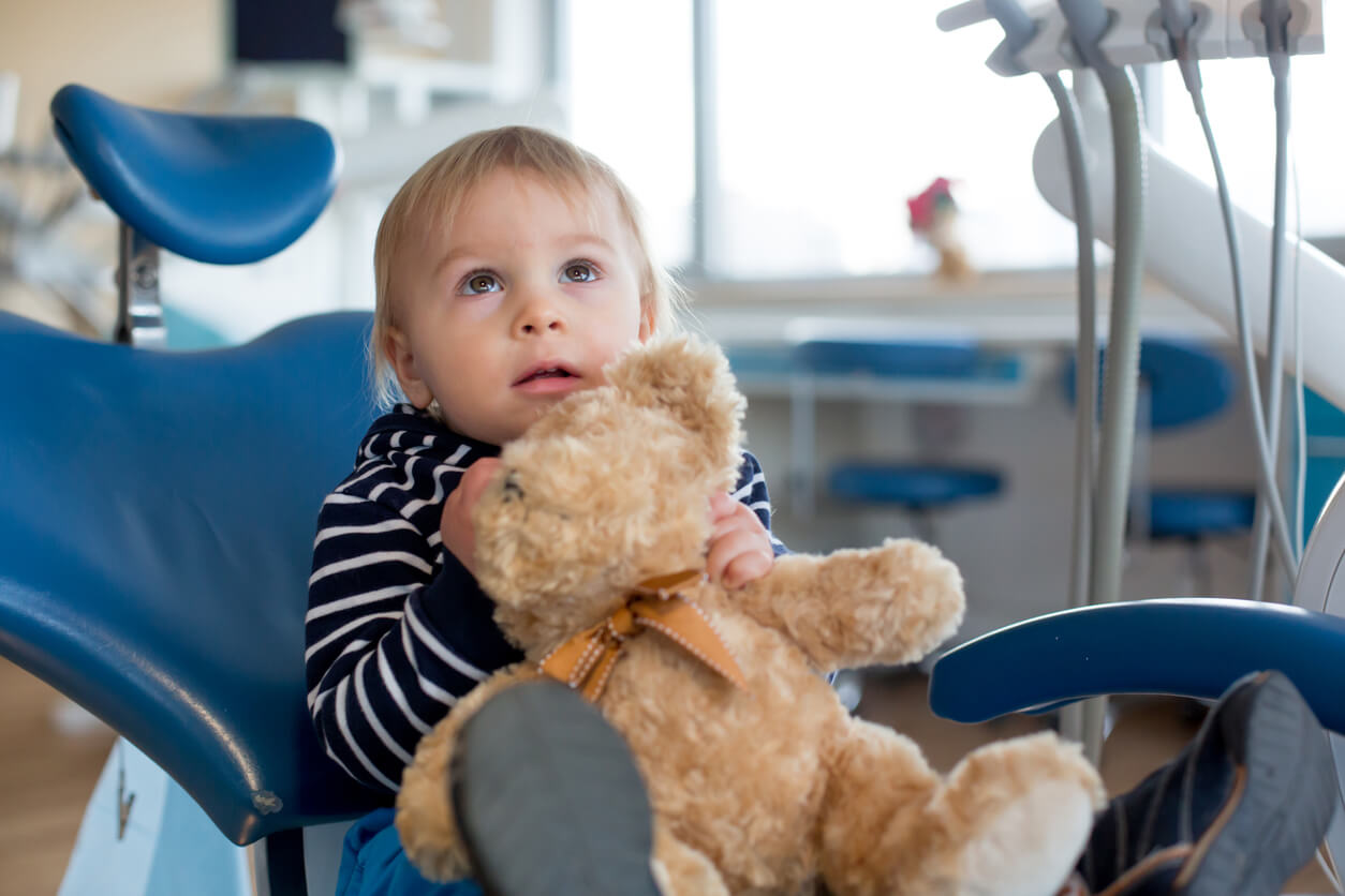 A toddler sitting in a dentist chair holding a teddy bear.