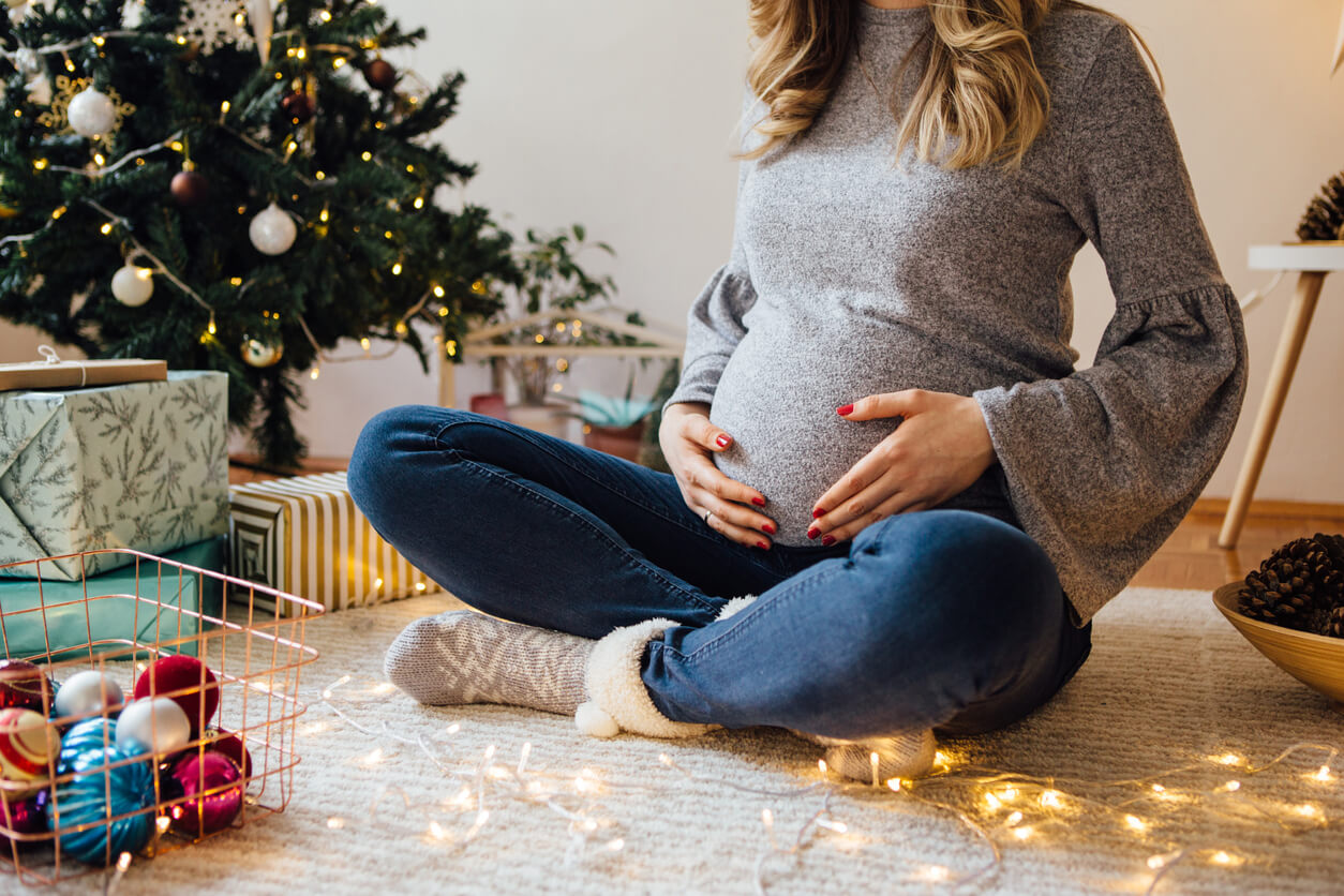 mujer embarazada sentada arbol navidad alfombra medias sweater vientre agarra amor regalos