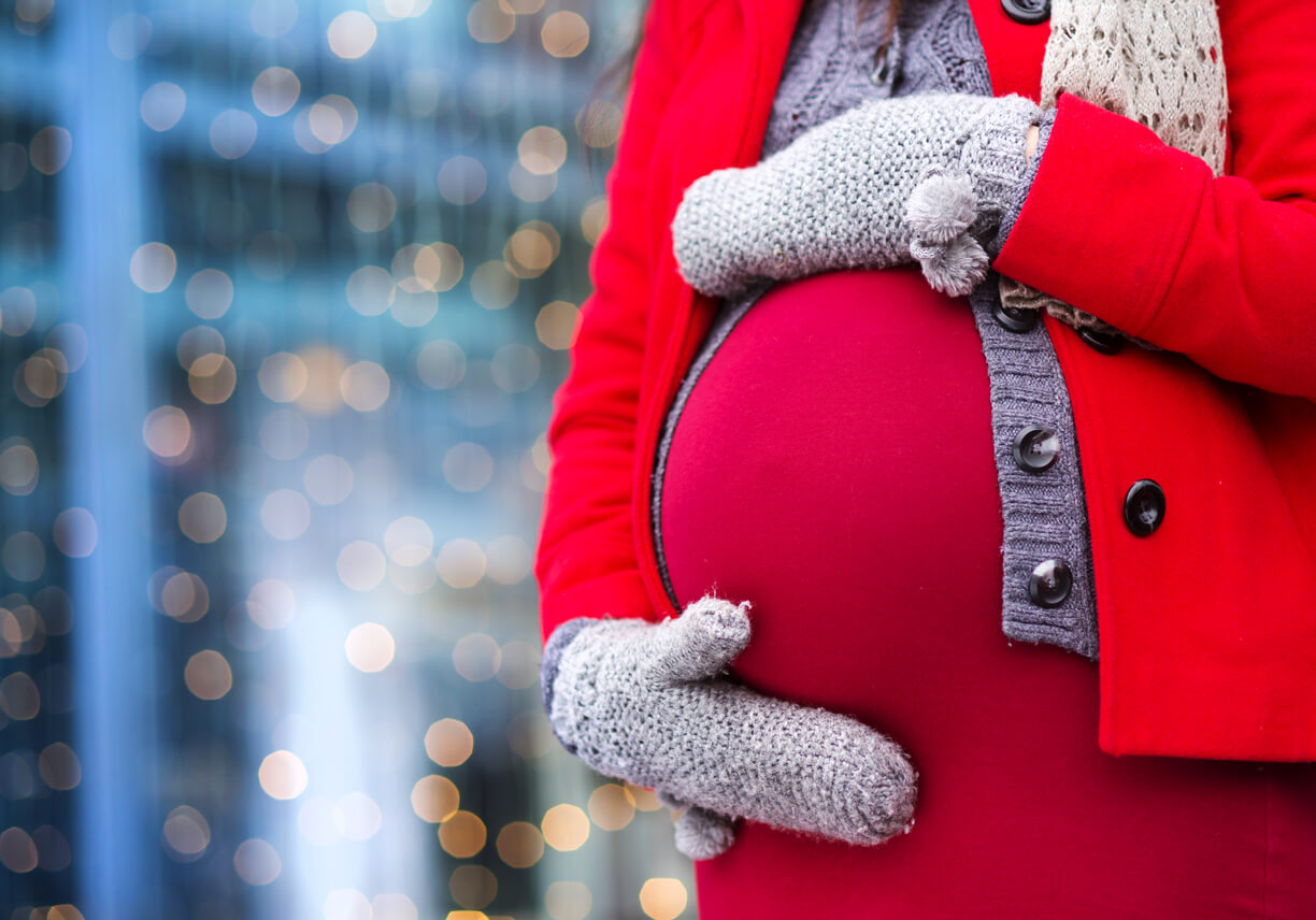 A pregnant woman wearing a red jacket and grey mittens.