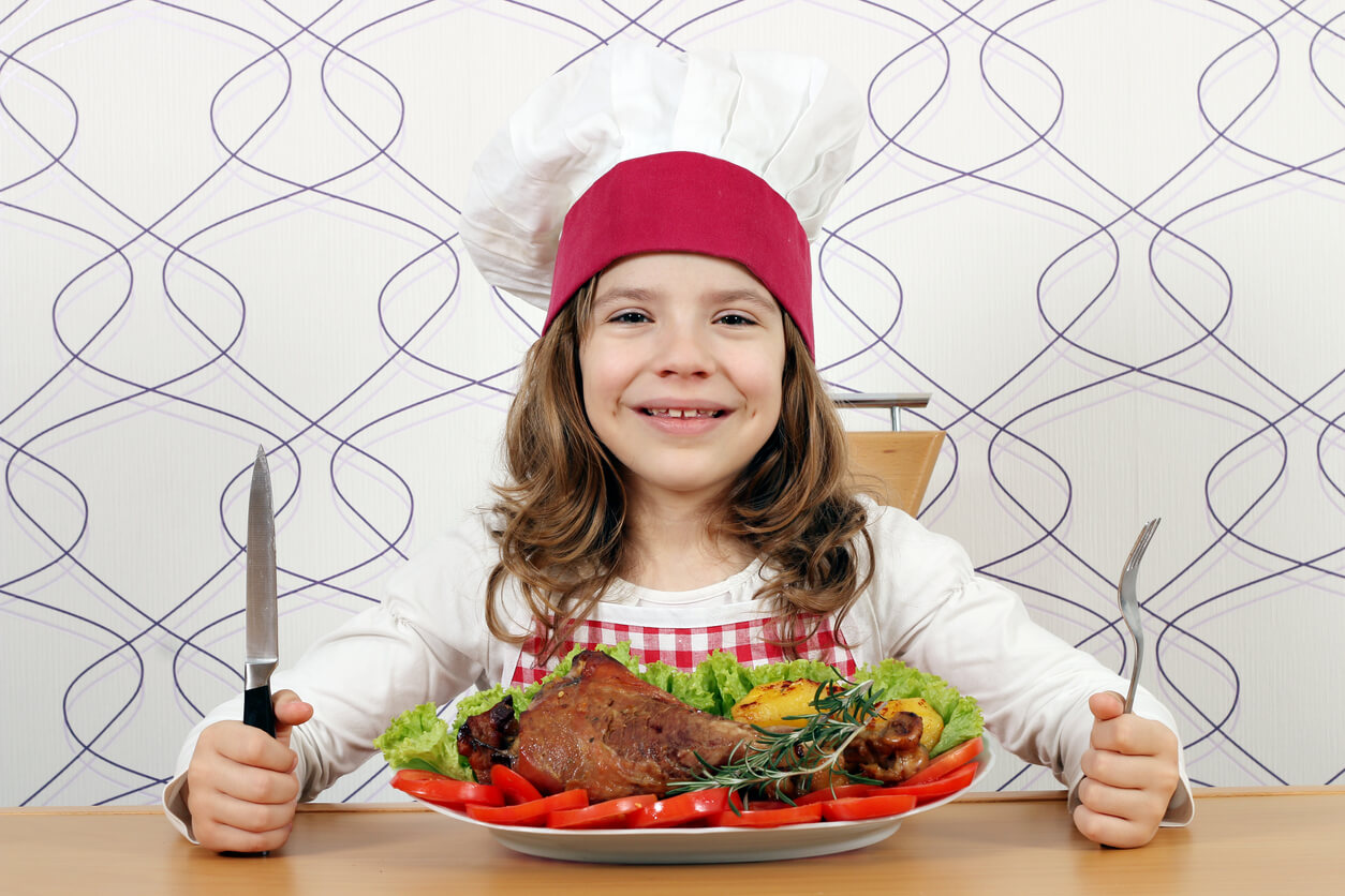 A girl wearing a chef hat and apron sitting down to eat a plate of meat and vegetables.
